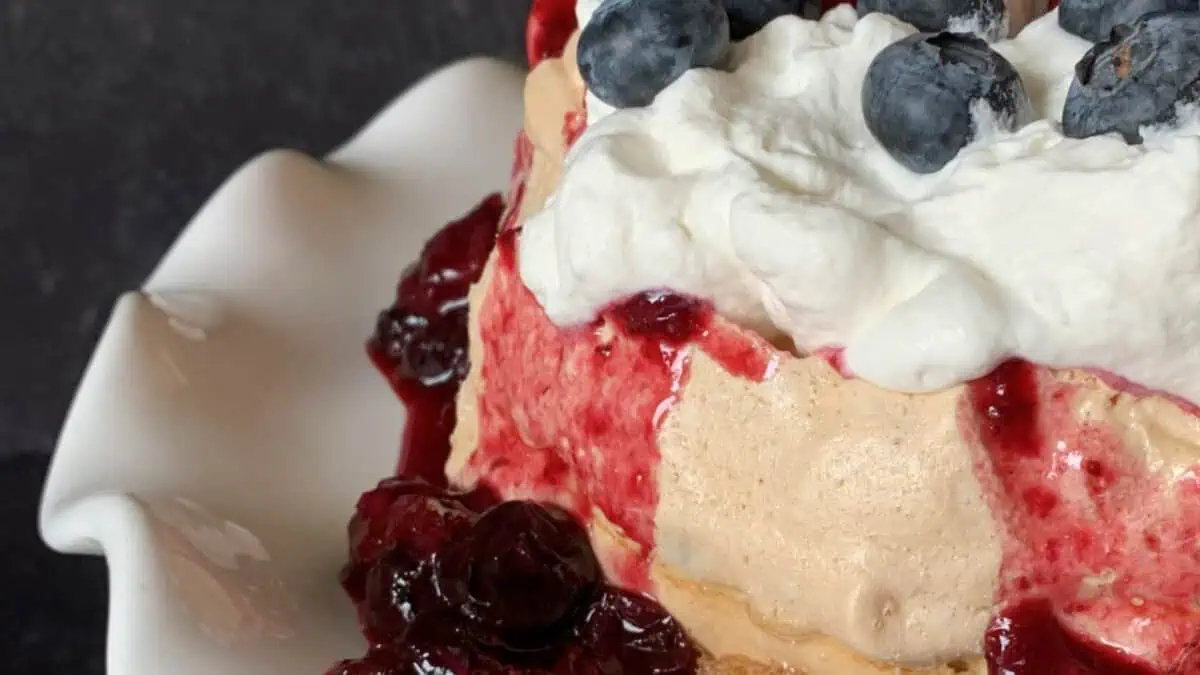 A close-up of a dessert featuring a creamy layered cake topped with whipped cream and blueberries. The cake is adorned with a rich red berry sauce that drips down its sides, sitting on a white scalloped dish. The background is dark, making the dessert stand out.