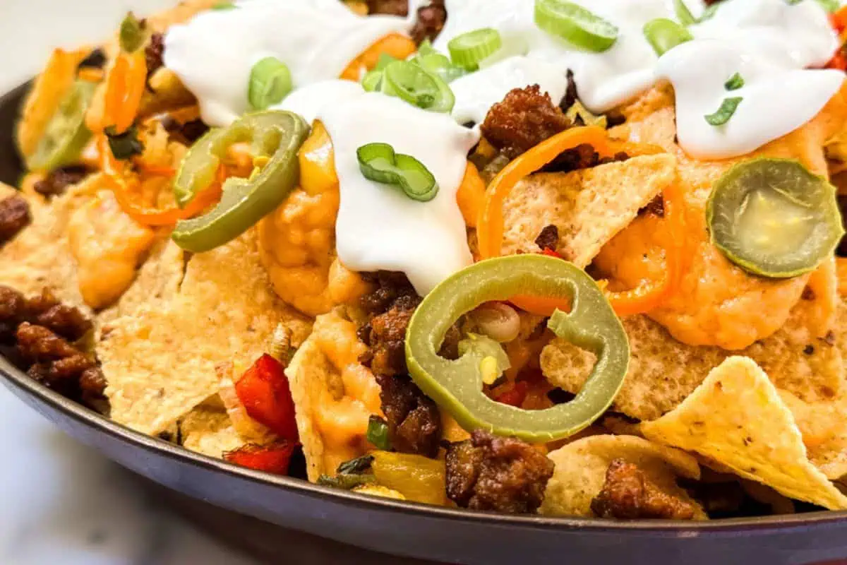 A close-up shot of a nachos dish piled high with tortilla chips, ground beef, melted cheese, jalapeño slices, sour cream, and chopped green onions. The vibrant toppings add color and texture to the dish.