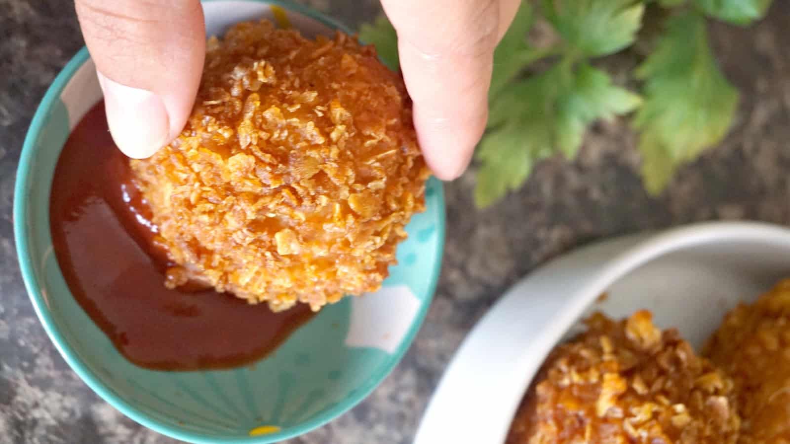 A hand holding a chicken ball to dip in ketchup.