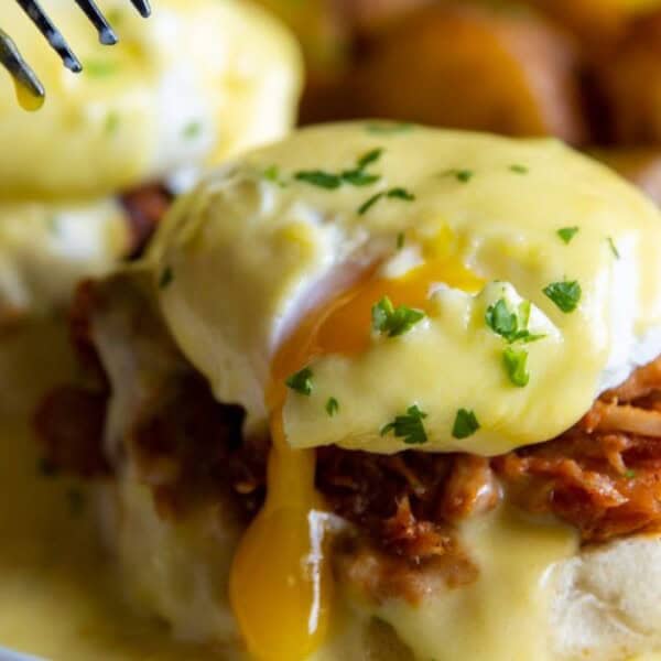 A close-up of eggs Benedict with pulled pork on an English muffin, topped with a poached egg and creamy hollandaise sauce. Garnished with chopped parsley, the yolk is oozing out. Roasted potatoes are visible in the background.