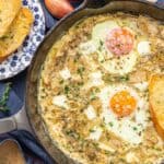 A skillet with baked eggs topped with fresh herbs, accompanied by crispy bread slices. A side plate holds additional bread.