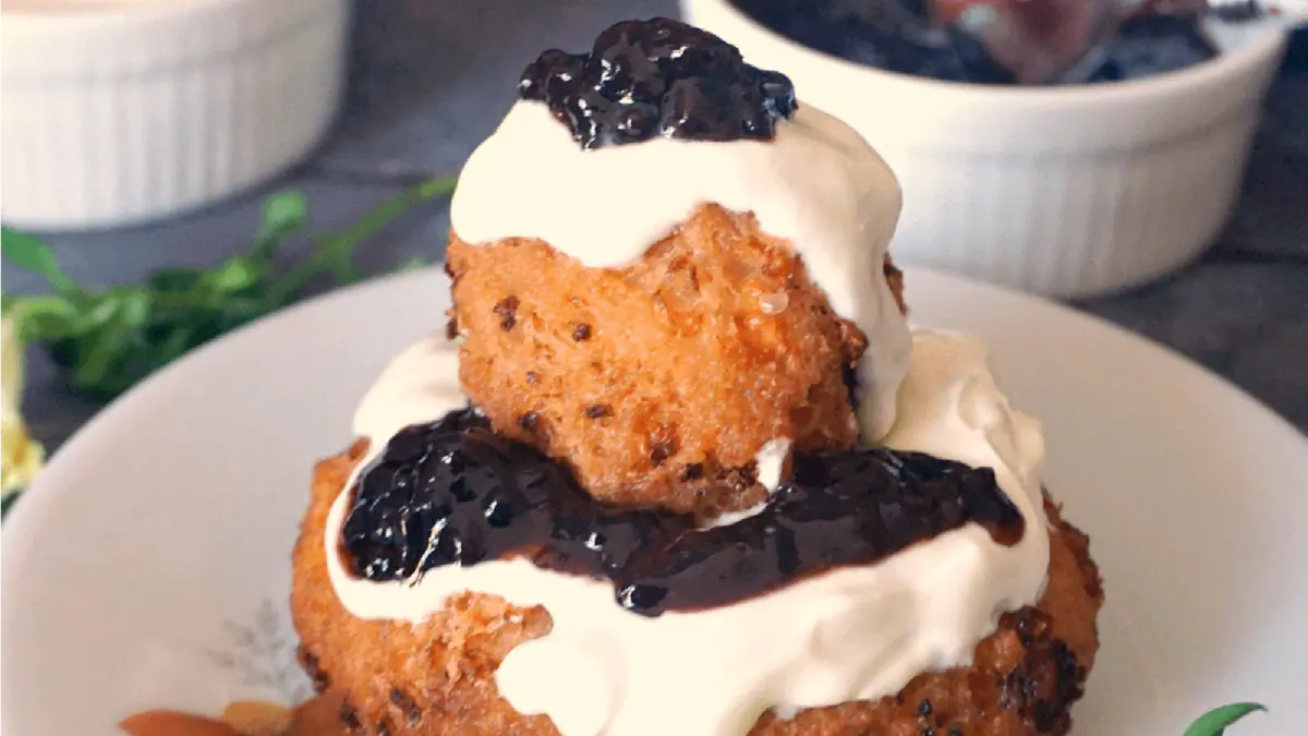 Two fried dough balls are stacked and topped with a generous dollop of cream or yogurt and a dark berry compote. The background includes white ramekins and hints of greenery, all set on a white plate.