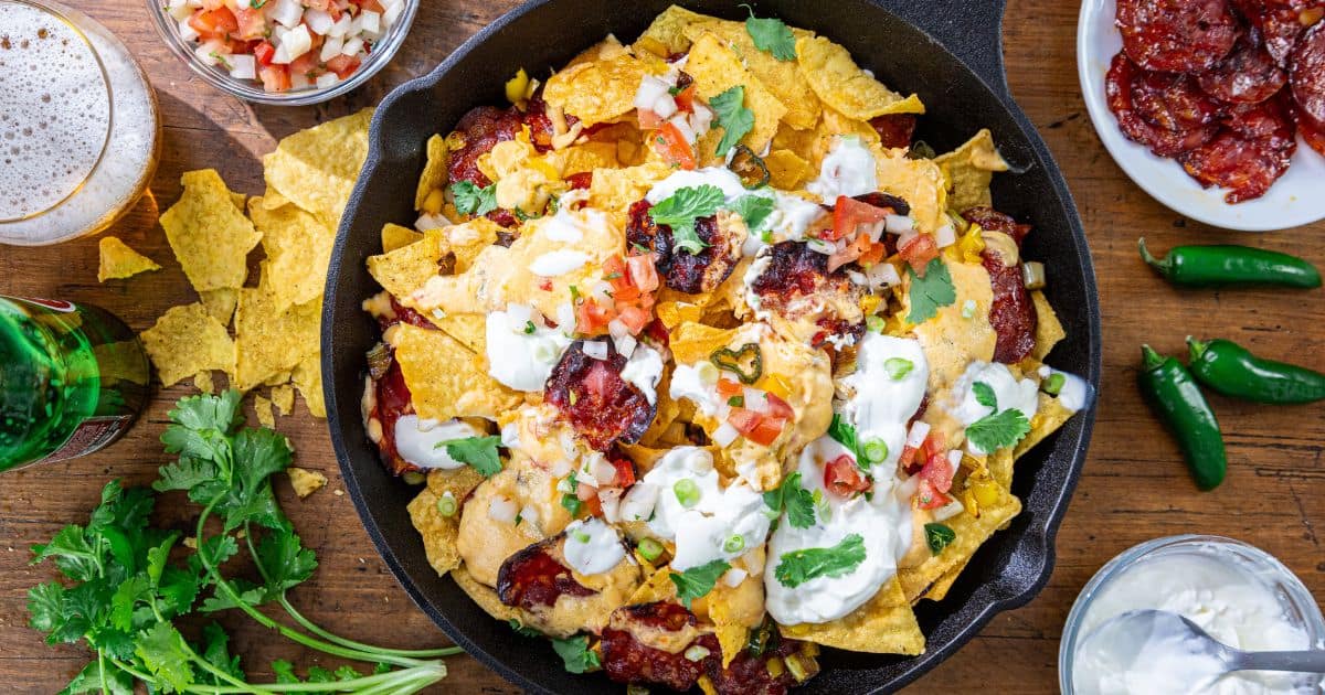 Pimento cheese nachos in a skillet with melted cheese, sour cream, salsa, chorizo, cilantro; jalapeños and dips nearby.