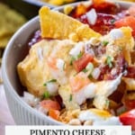A bowl of pimento cheese nachos topped with chopped onions, tomatoes, and parsley; chips are in the background.