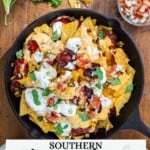 A skillet of pimento cheese nachos with sour cream, cilantro, and salsa sits on a wooden table next to extra salsa and sour cream.