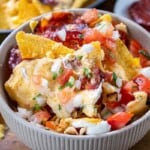 A bowl of nachos with melted cheese, chopped tomatoes, onions, cilantro, and slices of cured sausage.