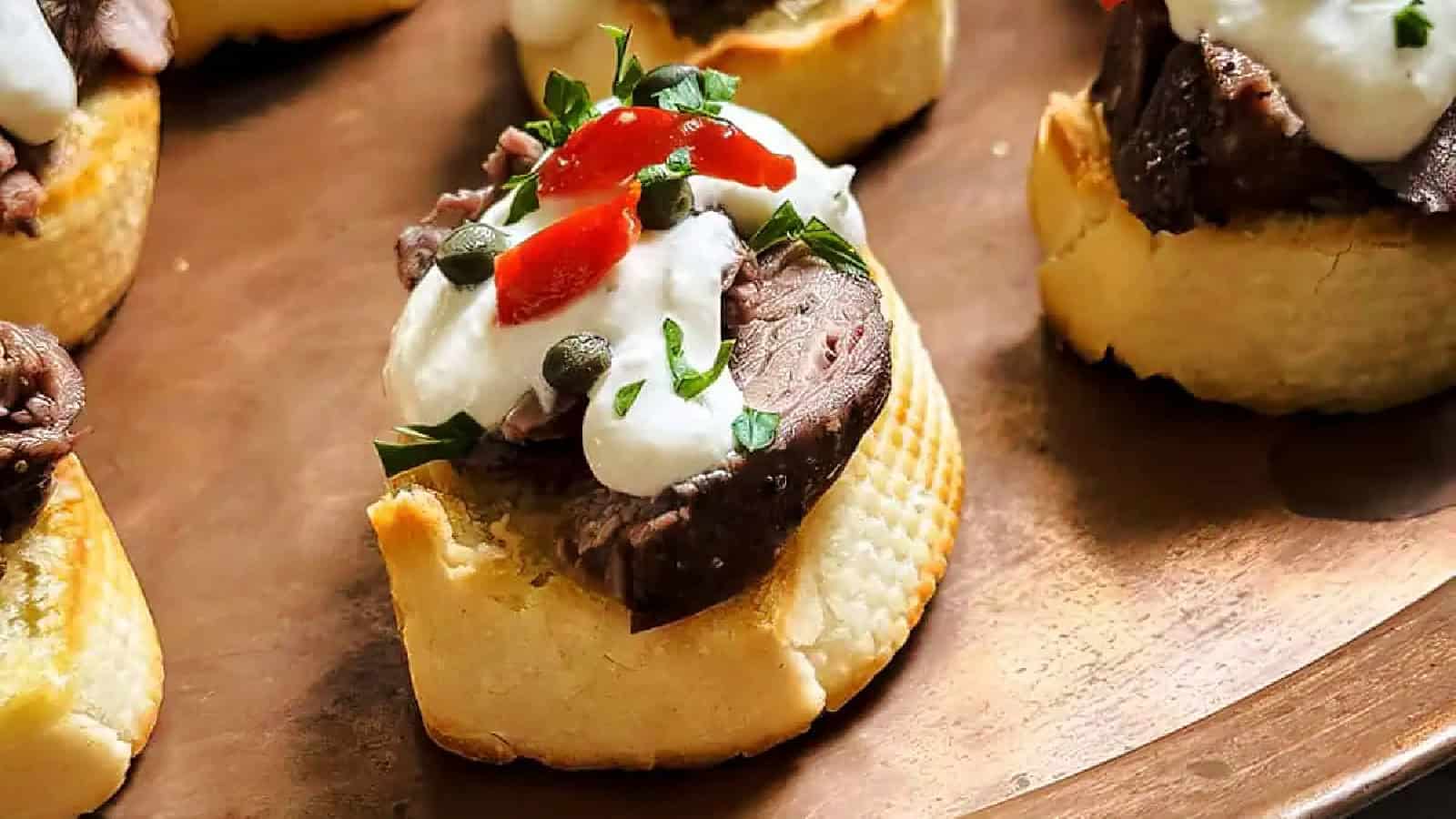Open-faced sandwiches with sliced roast beef, creamy sauce, red peppers, and capers on a parchment-lined surface.