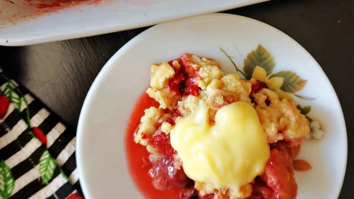 A white plate holds a serving of mixed berry cobbler topped with a dollop of pale yellow custard. The cobbler features a crumbly golden crust with visible red berry filling. Part of a colorful, patterned cloth is seen to the left of the plate.