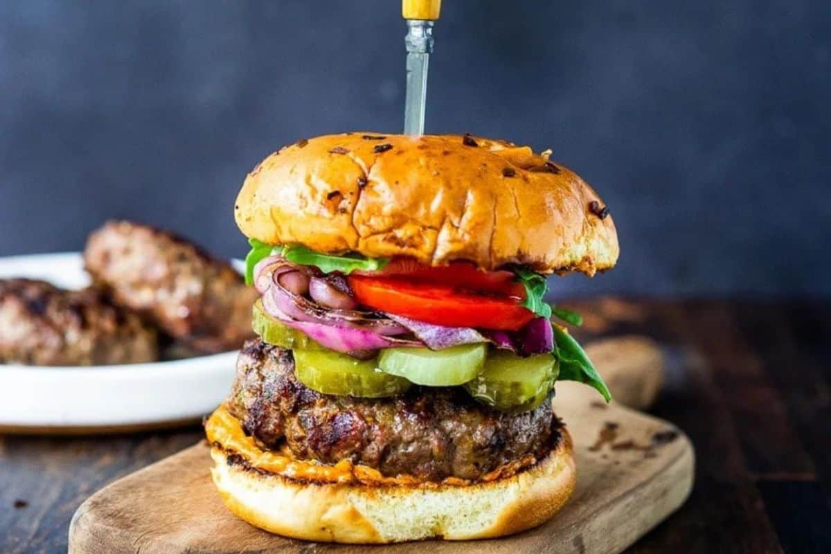 A grilled burger with lettuce, tomato, pickles, onions, and sauce on a toasted bun is pierced by a knife and set on a wooden board.