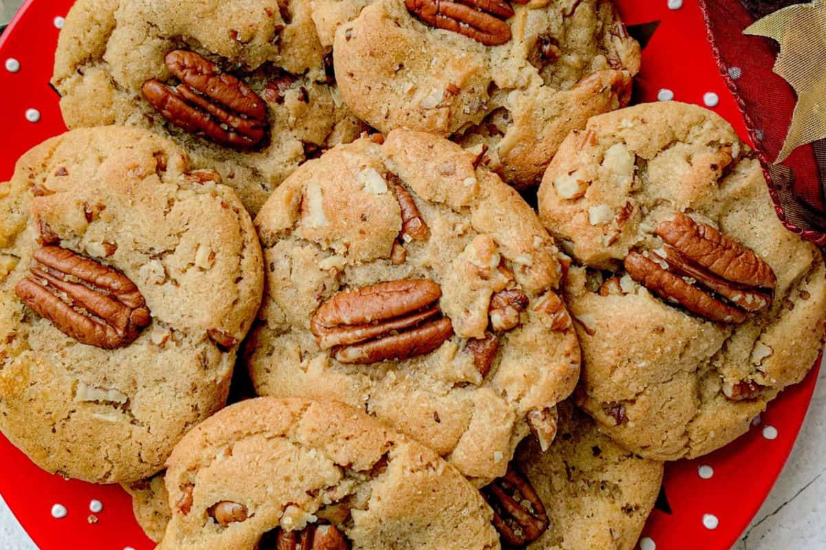 A red plate with white polka dots holds several cookies. Each cookie is topped with a whole pecan, and there are scattered pecan pieces on the cookies.