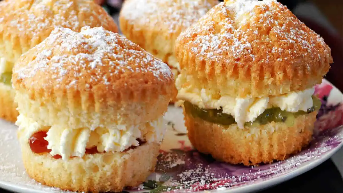Four scones filled with cream and jam are displayed on a floral-patterned plate. The scones are topped with a light dusting of powdered sugar.