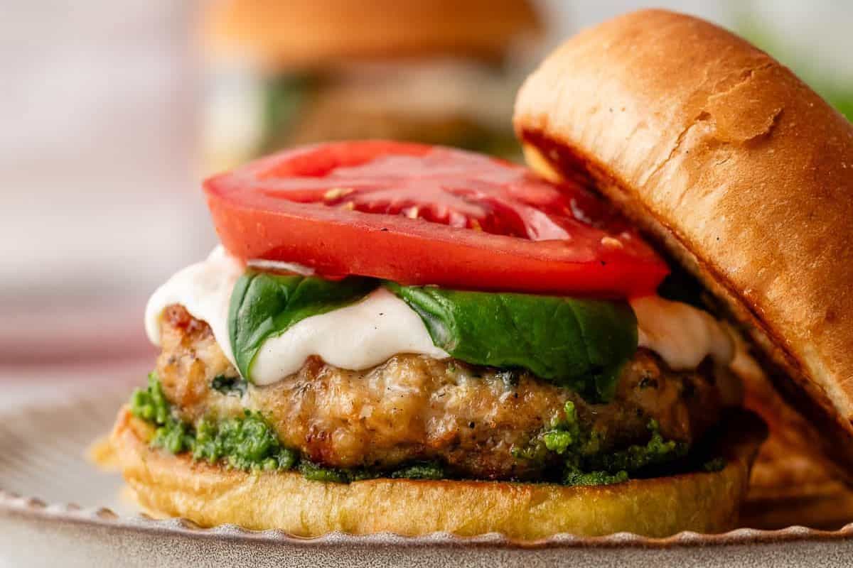 A grilled burger with a bread bun, grilled patty, green pesto, spinach leaves, melted cheese, and two slices of tomato on a plate.