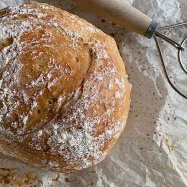 A round loaf of bread dusted with flour beside a whisk.