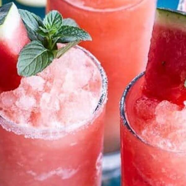 Four glasses filled with pink slushy drinks, each garnished with a watermelon wedge and mint leaves, arranged on a blue surface.