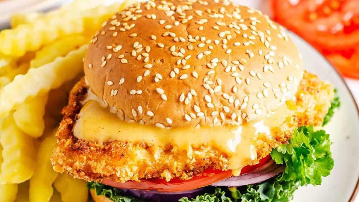 A breaded chicken sandwich on a sesame seed bun, topped with lettuce, tomato, onion, cheese, and sauce, served with crinkle-cut fries.