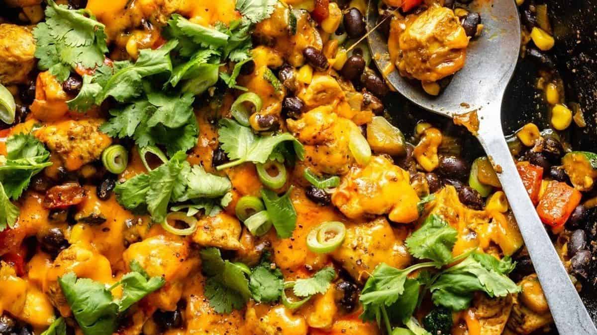 A skillet filled with cheesy Mexican chicken, black beans, corn, red bell peppers, and chopped cilantro. A large spoon is placed beside the skillet.