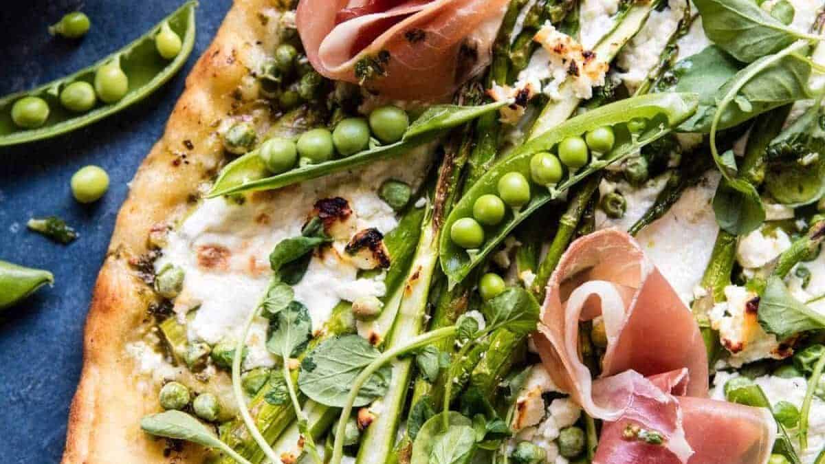 Flatbread topped with asparagus, sugar snap peas, prosciutto, crumbled cheese, and fresh greens. Served on a dark blue surface.