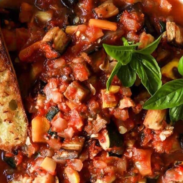 A bowl of chunky vegetable stew with zucchini and squash, topped with fresh basil, next to a slice of toasted bread.
