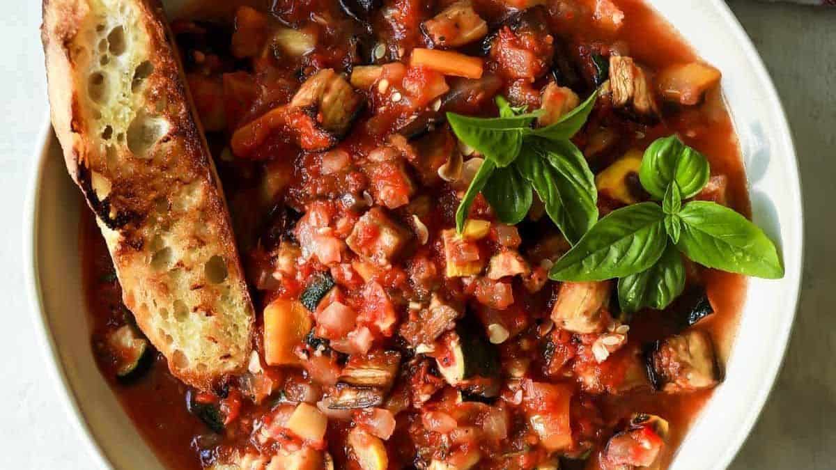 A bowl of chunky vegetable stew with zucchini and squash, topped with fresh basil, next to a slice of toasted bread.