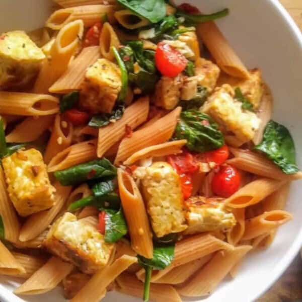 Large white bowl of lentil pasta, tempeh pieces, spinach, and tomatoes on wood cutting board and napkin with loaf of bread next to it.