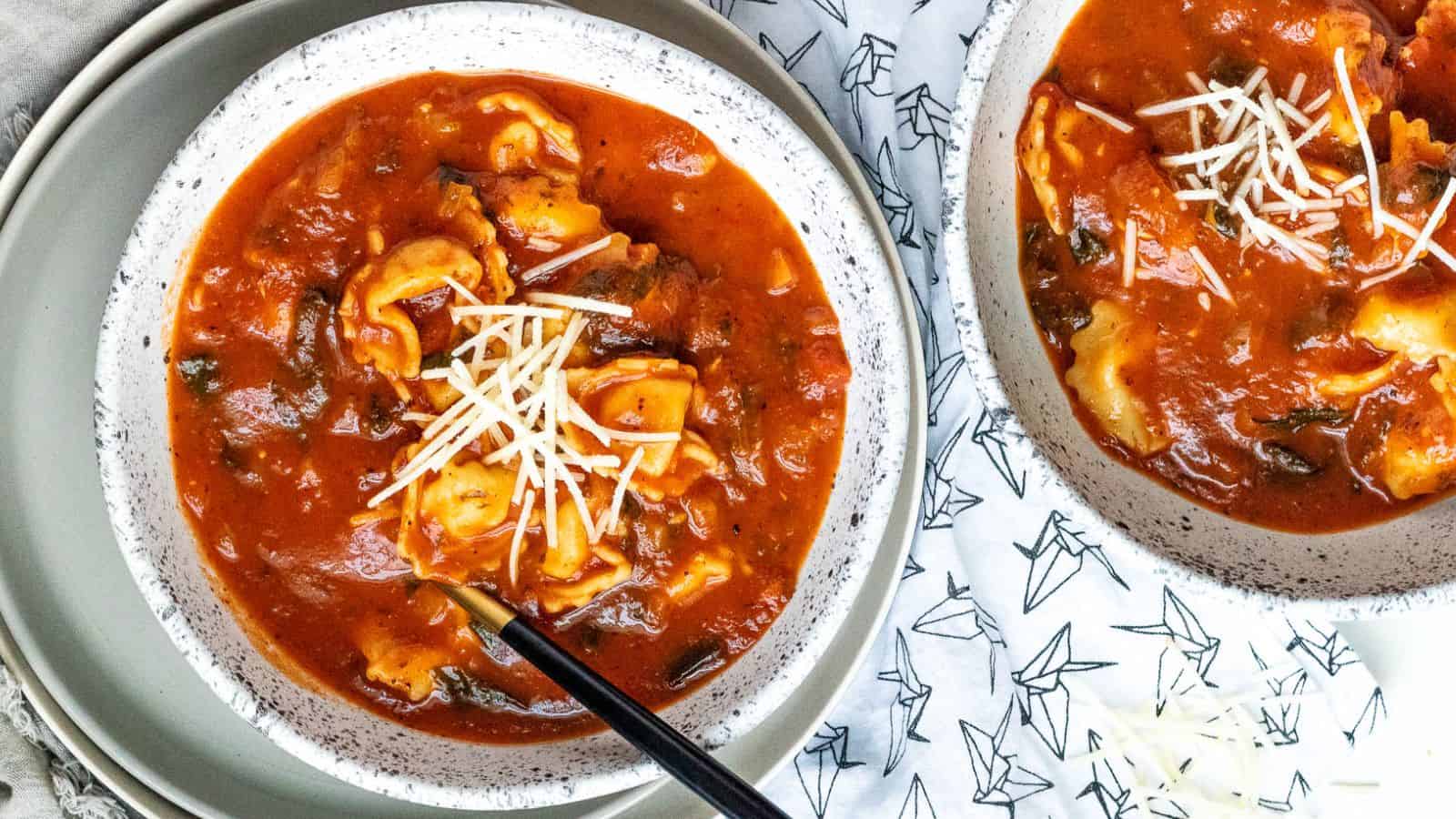 Two bowls of tomato soup with tortellini and shredded cheese. A spoon rests in one bowl.