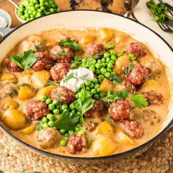 A Dutch oven containing a stew of meatballs and potatoes with peas and gravy and a dollop of sour cream.