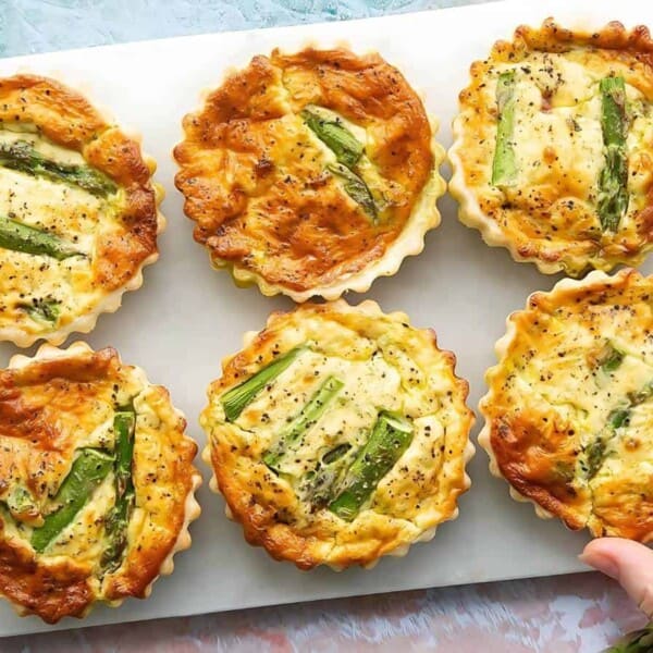 Six small asparagus quiches on a white cutting board, with a hand touching one. Cherry tomatoes and a green condiment in the background.