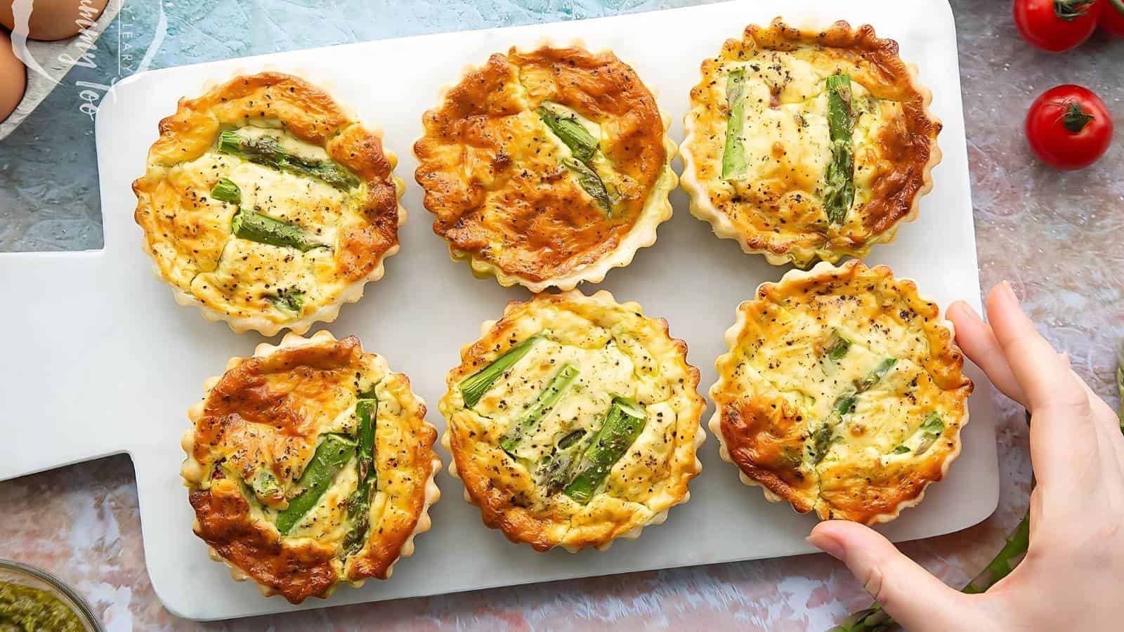 Six small asparagus quiches are arranged on a white cutting board. The golden-brown crusts are visible, with asparagus spears on top. A hand is reaching to pick one up. There are ripe tomatoes in the background.