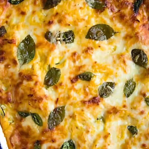A freshly baked lasagna in a rectangular dish, topped with melted cheese and basil leaves. A hand is gripping the side of the dish.