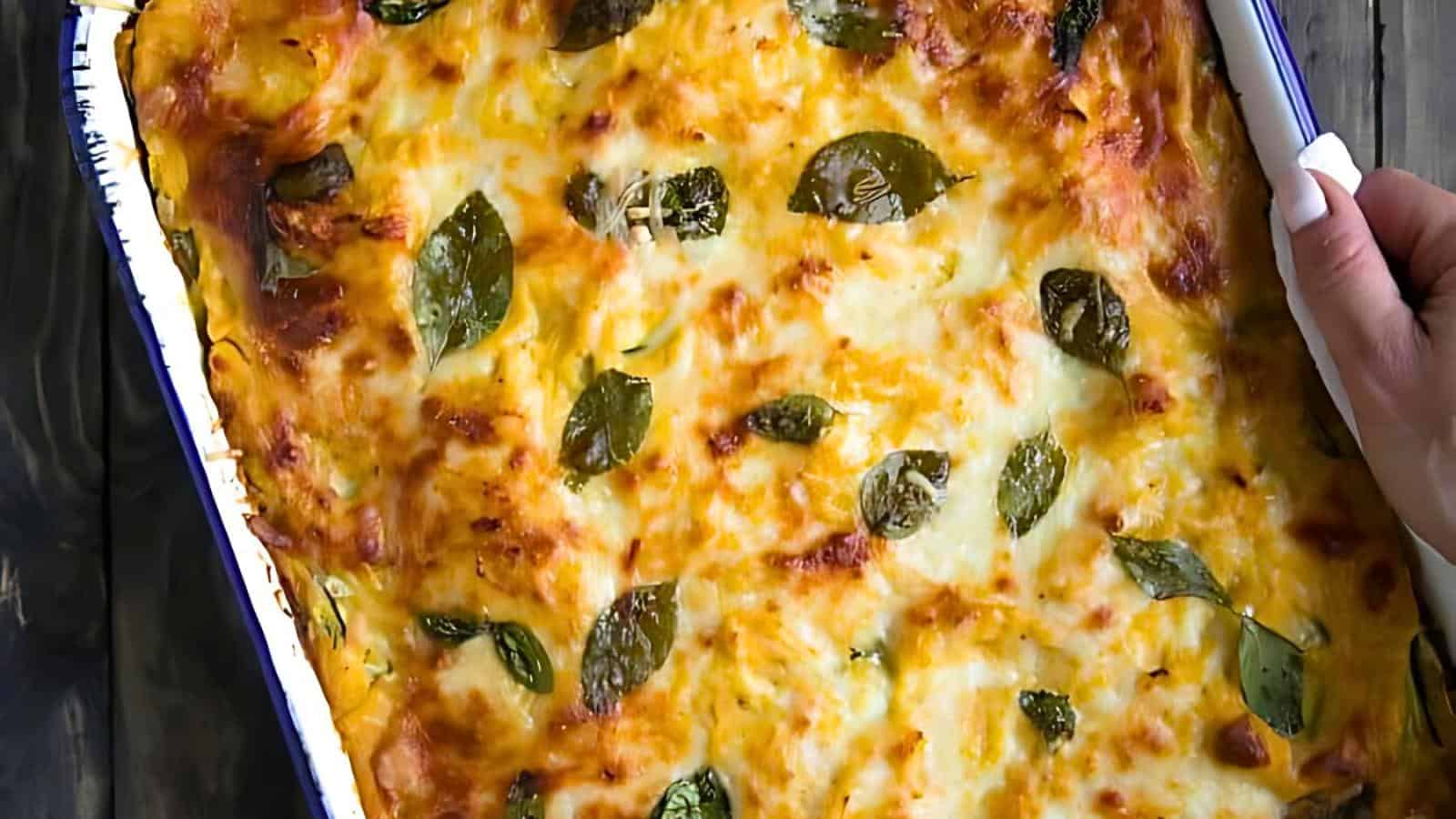 A freshly baked lasagna in a rectangular dish, topped with melted cheese and basil leaves. A hand is gripping the side of the dish.