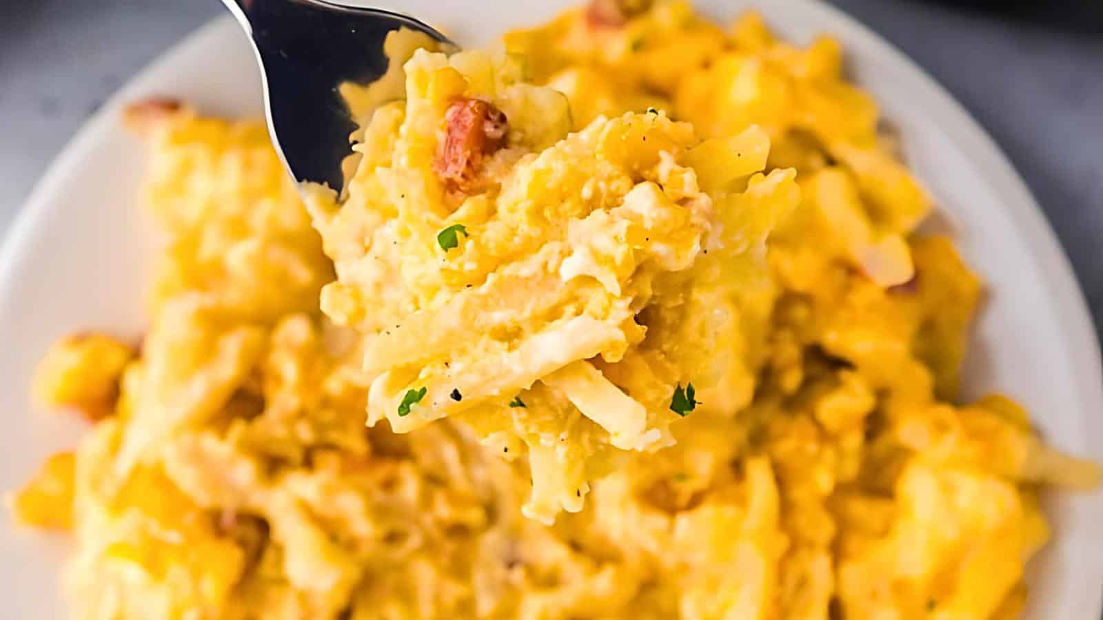 A fork holding a bite of cheesy hash brown casserole with ham, with a plate of the same dish in the background.
