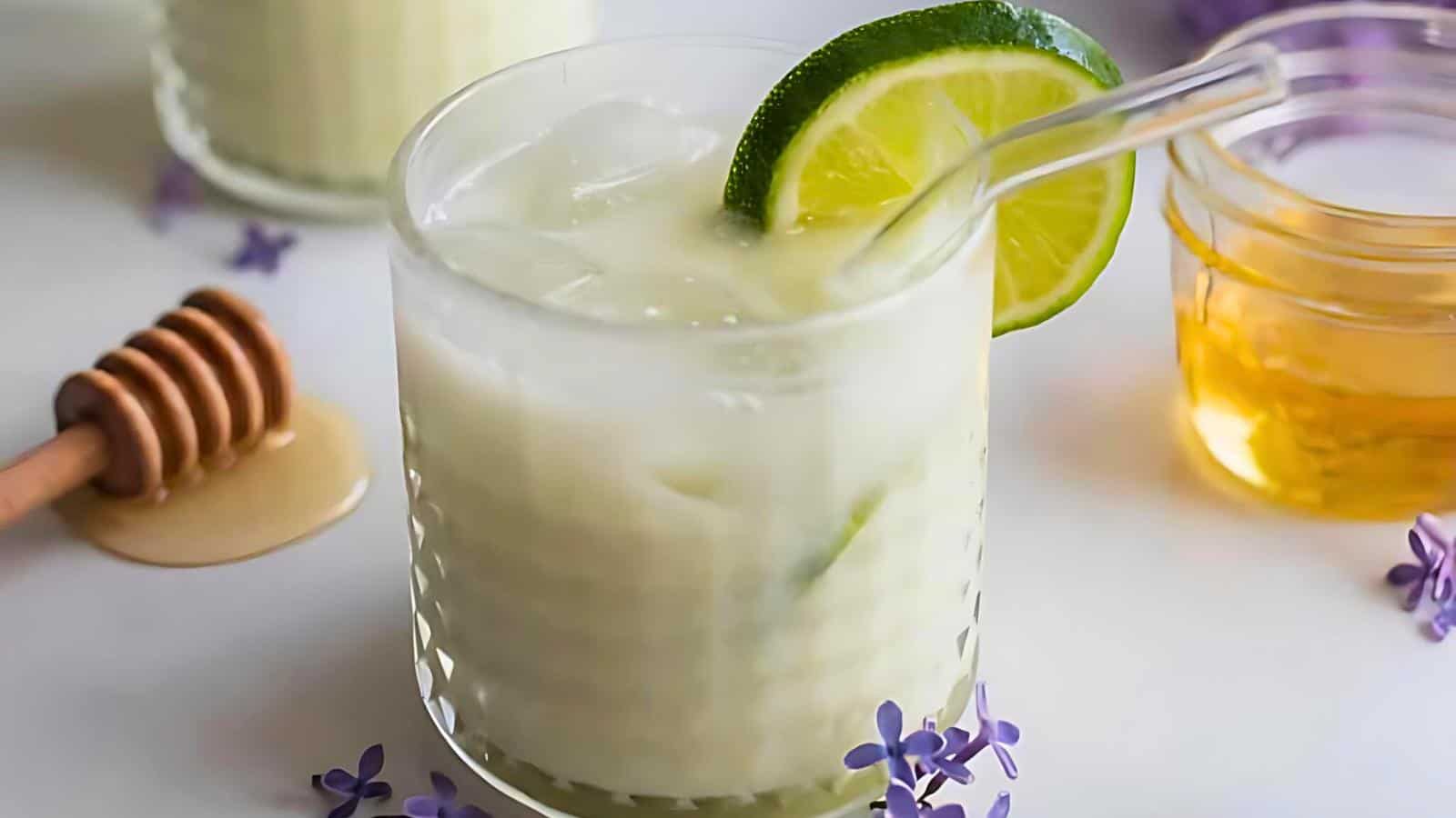A glass of iced drink with a lime slice and a glass straw, next to a jar of honey and a honey dipper. Small purple flowers scattered on a white surface.