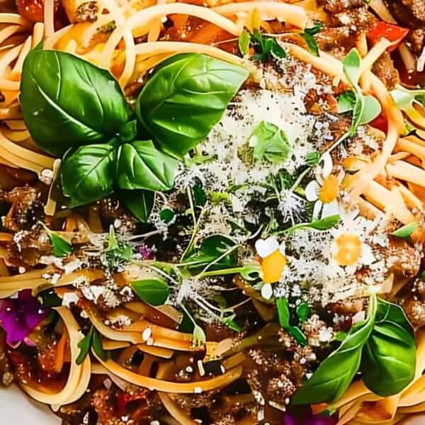 A plate of spaghetti with meat sauce, fresh basil leaves, grated cheese, edible flowers, and a fork to the side.