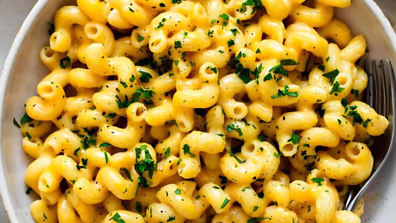 A close-up image of a bowl filled with creamy macaroni and cheese. The elbow pasta is coated in a rich, yellow cheese sauce and garnished with finely chopped green herbs and a sprinkling of black pepper. A fork is placed on the side of the bowl.