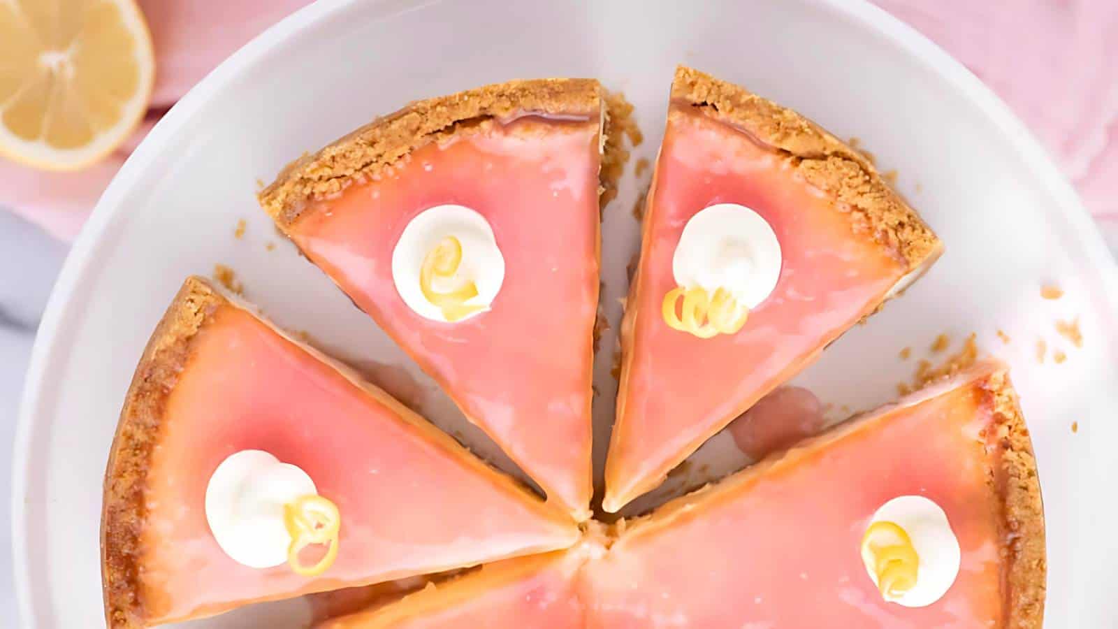 A round lemon-pink pie cut into six slices, topped with dollops of whipped cream and lemon twists, displayed on a white plate.