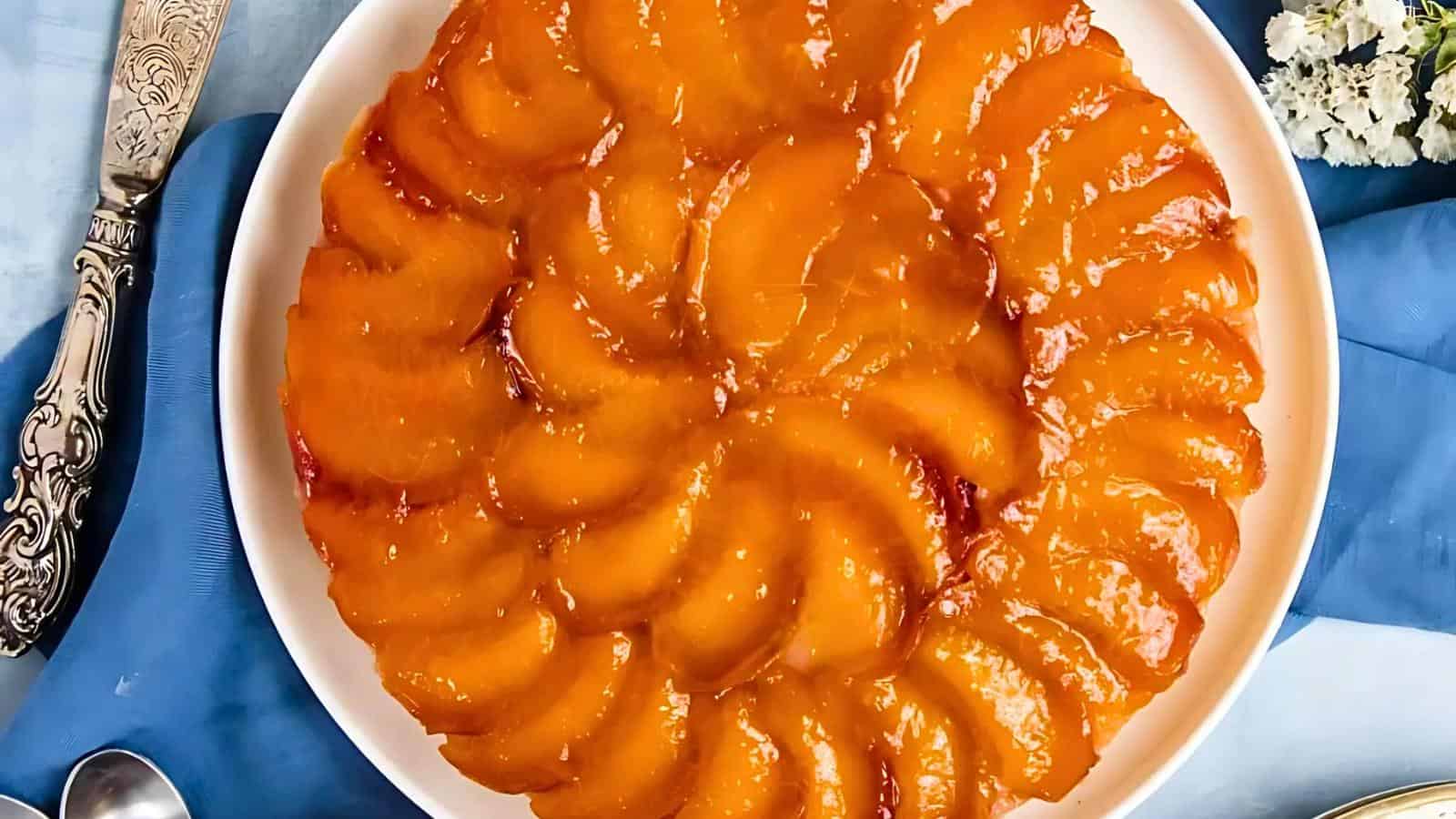 A close-up photo of a peach tart arranged in a circular pattern on a white plate. The glistening peach slices are neatly aligned, creating a visually appealing design. The plate rests on a blue cloth, and there are decorative silver utensils nearby.