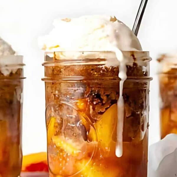 Three glass jars filled with peach cobbler, topped with melting vanilla ice cream and accompanied by a spoon. The cobbler layers show peach slices and a brown crumble.