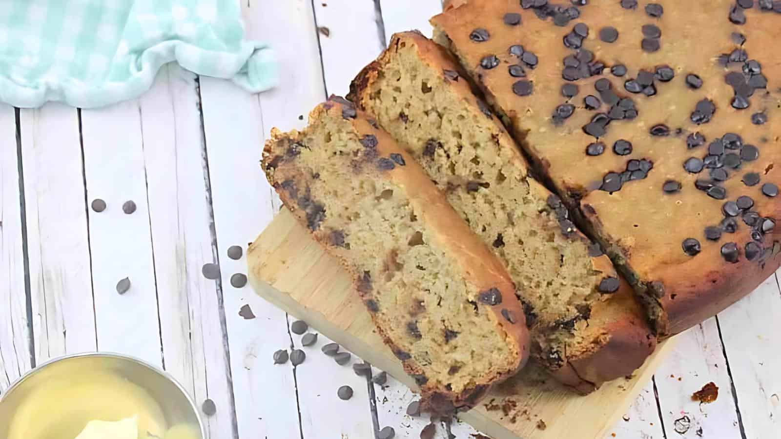 A sliced loaf of chocolate chip banana bread rests on a wooden cutting board. In the background, there is a light blue checkered cloth and a bowl of butter.