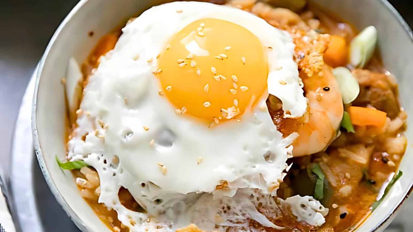 A bowl of rice topped with a fried egg, assorted vegetables, shrimp, carrots, greens, and sprinkled sesame seeds.