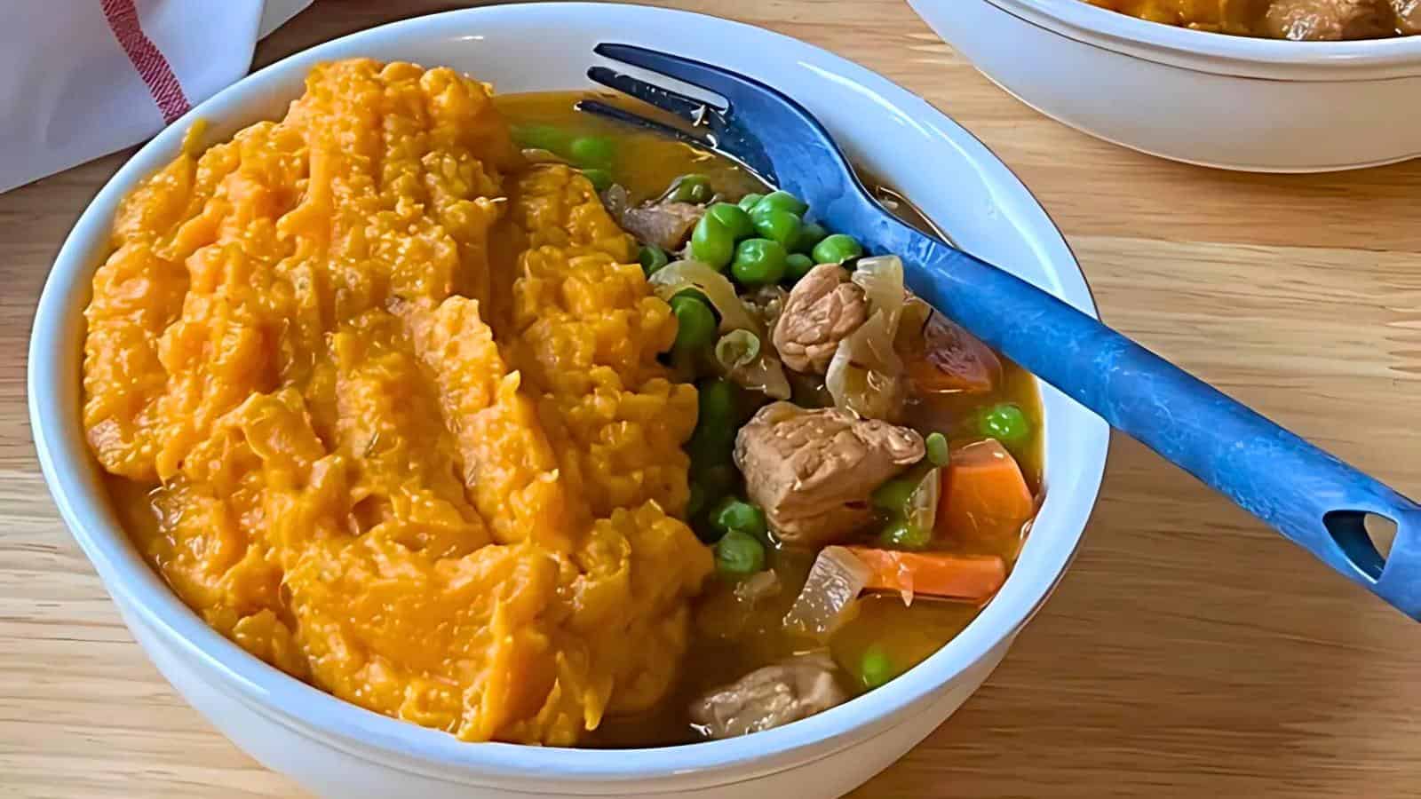 A blue fork alongside a bowl containing mashed sweet potatoes, cooked peas, chopped carrots, and chunks of meat on a wooden table.
