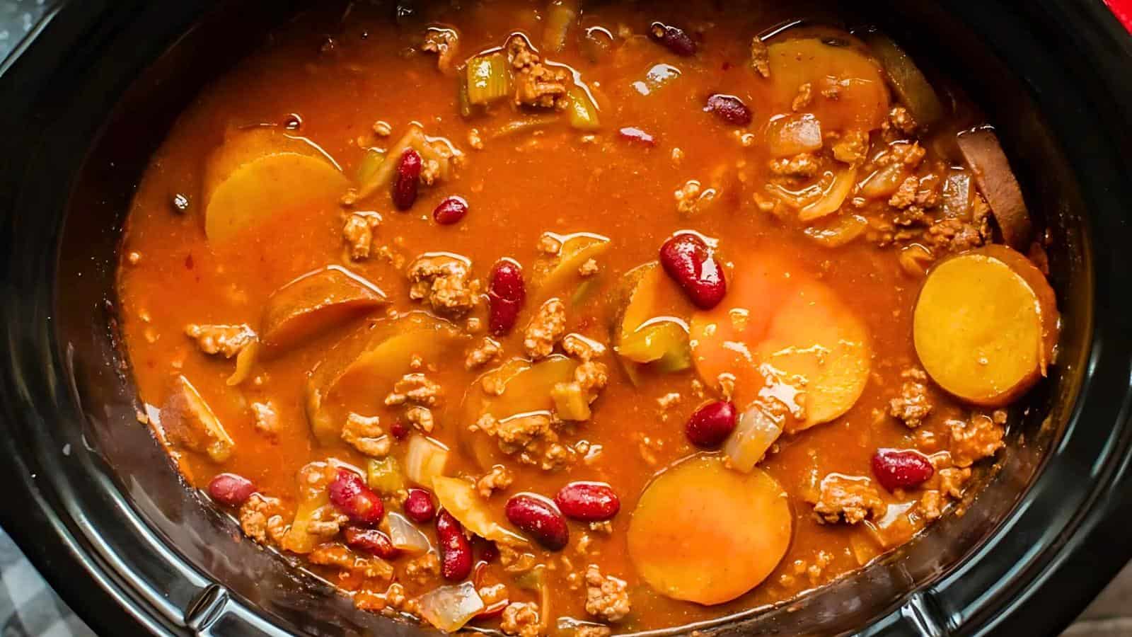 A slow cooker filled with stew containing ground meat, kidney beans, sliced potatoes, and vegetables in a red-brown broth.