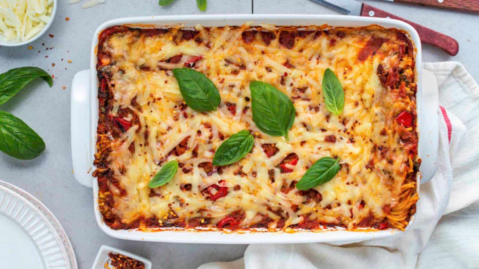 A baked casserole dish filled with cheesy lasagna, topped with melted cheese and fresh basil leaves. The dish is placed on a light-colored surface with a cloth napkin and some scattered ingredients nearby, including shredded cheese and basil.