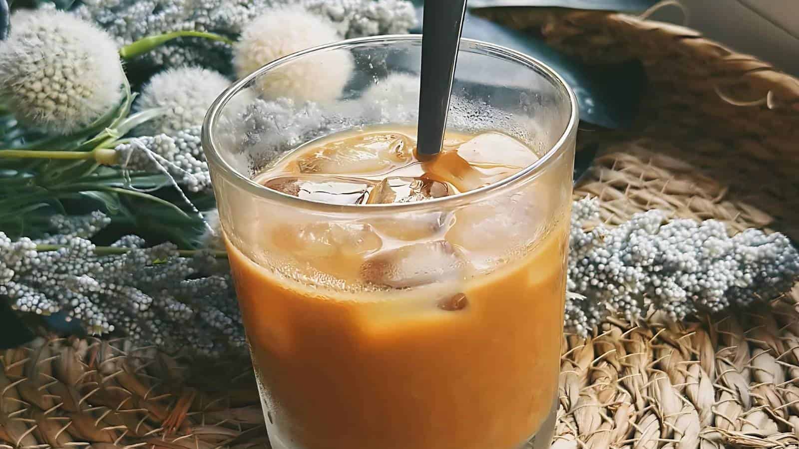 A glass of iced coffee with a straw, placed on a woven mat, with white flowers in the background.