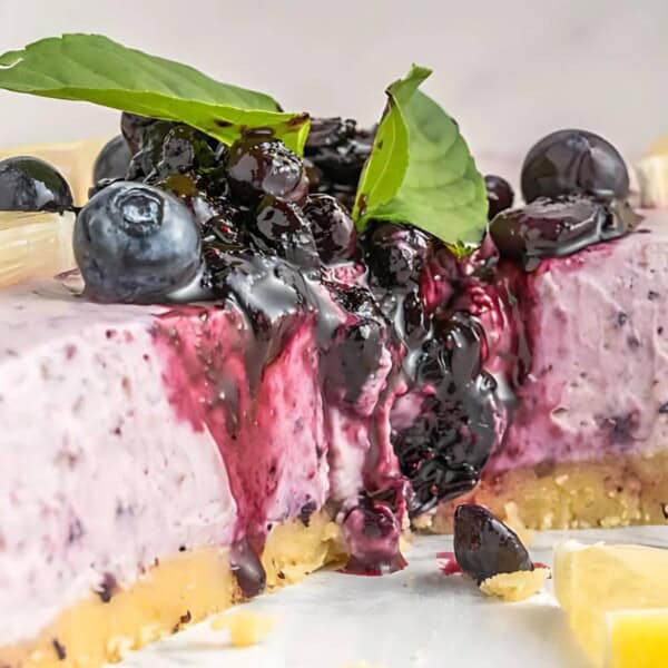A blueberry cheesecake with fresh blueberries, lemon slices, and mint leaves on top. The cheesecake is partially sliced, showing a creamy, purple interior and a crumbly crust.