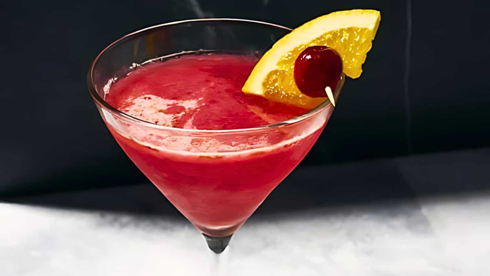 A red cocktail in a martini glass garnished with an orange slice and a cherry on a toothpick.
