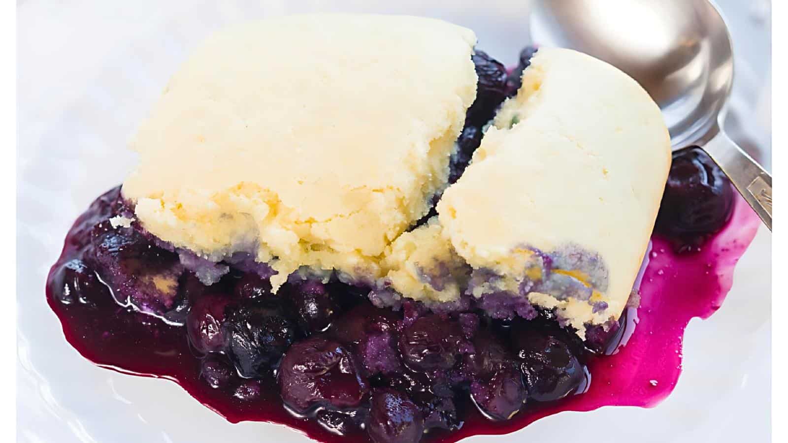 A serving of blueberry cobbler with a golden crust, juicy blueberries beneath, on a white plate with a spoon.