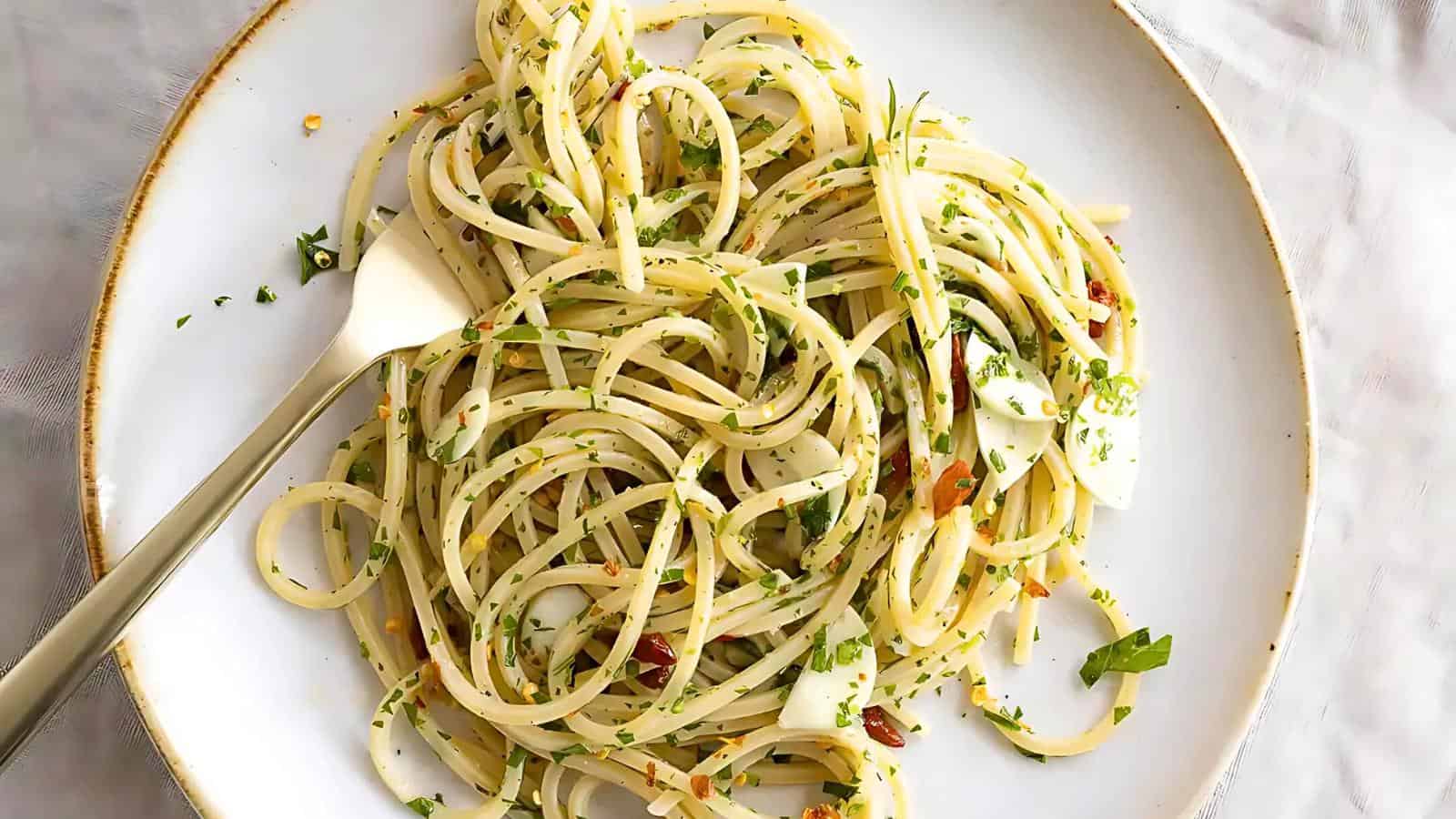 A white plate with a serving of spaghetti aglio e olio, garnished with chopped parsley and red pepper flakes. A fork is twirled into the pasta, and thin slices of garlic are visible among the noodles.