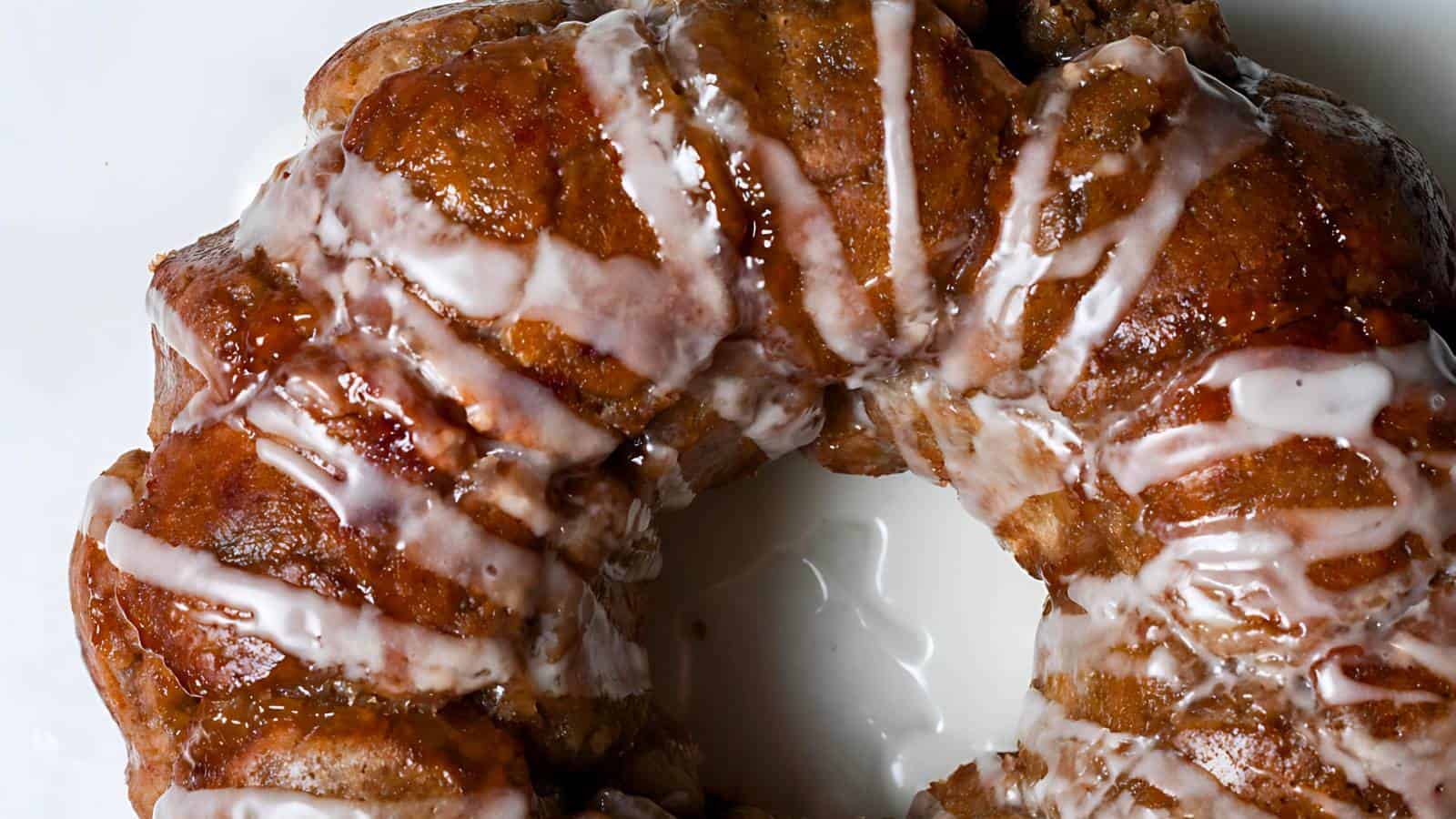 A close-up of a cinnamon monkey bread ring drizzled with white icing on a white plate. The bread's texture is rich and caramelized, highlighting its sweet and sticky appearance.