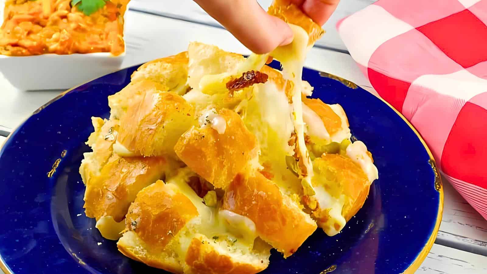 A hand pulling apart a cheesy, garlic-stuffed bread loaf on a dark blue plate. The melted cheese stretches as a piece is lifted. A small dish with dip is in the background on a white table with a red checkered cloth nearby.