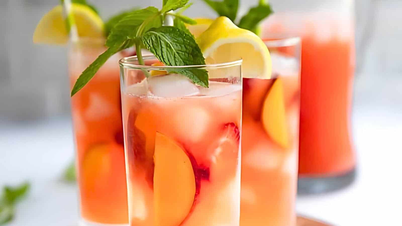 Three glasses of peach lemonade with mint leaves, lemon slices, and ice cubes, along with a pitcher of the same drink in the background.